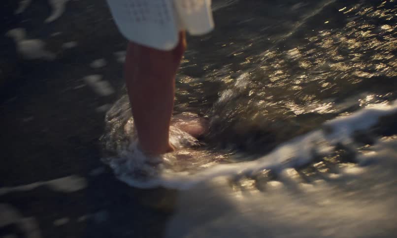 A person's bare feet stand in the wet sand of a beach shoreline at dusk with water and foam swirling around their ankles as the ocean waves come in and out in the dim light.