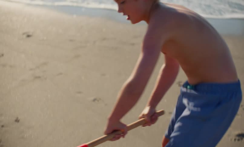 A young, shirtless blonde boy in blue shorts digs in the sand with a red plastic shovel near the shoreline on a sunny day at the beach as another kid plays nearby.