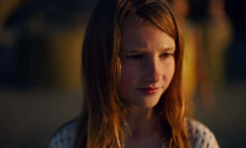 Portrait of a young red-headed girl with a thoughtful look on her face standing outdoors at sunset as the camera pans away revealing the blurred figure of another girl in a yellow dress standing behind her.