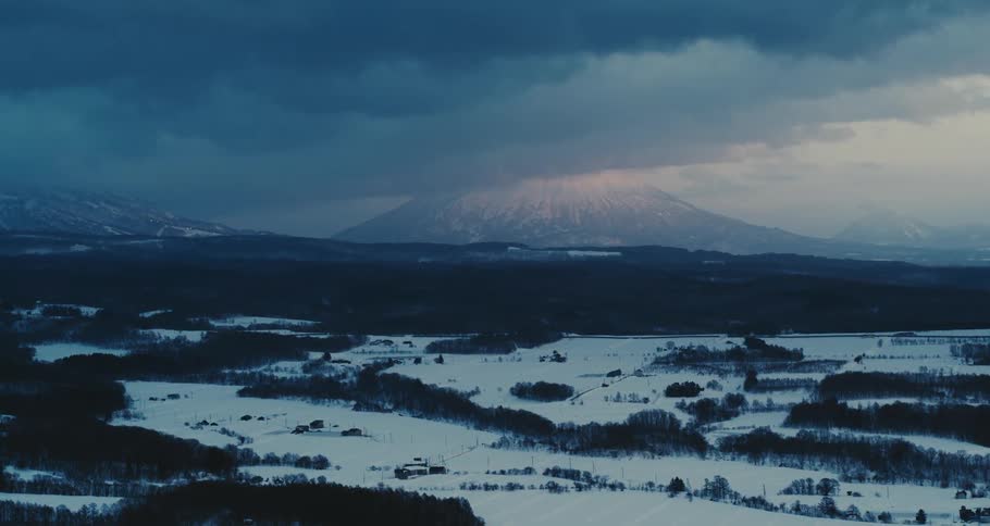 A snowy rural landscape stretches at dusk with a misty mountain in the background