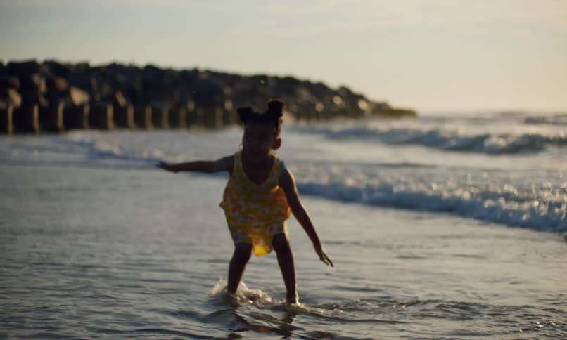 Slow motion of two diverse, happy little girls in sundresses who are excitedly splashing, kicking, and raising their arms at the surf and waves rolling in on the beach at sunset.