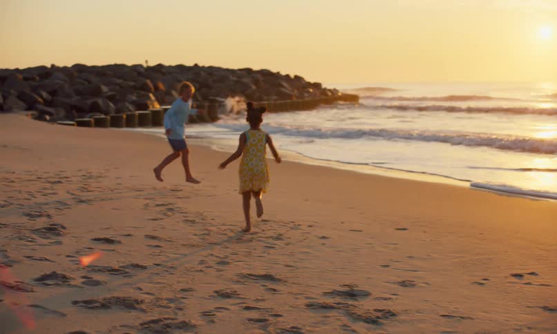 Three children running around and chasing each other at the beach under the golden rays of the setting sun as waves gently roll the surf on to the sand.