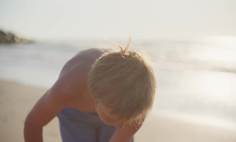 A little boy and girl work together on the beach to shovel sand into a plastic mold and pat it flat to make a sandcastle as the evening sun reflects off the surf coming in behind them.