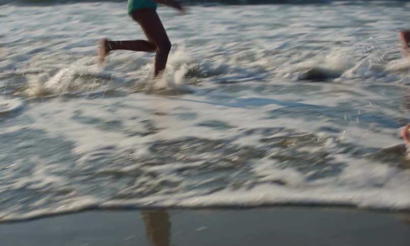 3 diverse children run and chase each other around in the waves and surf of a beach as the sun goes down.