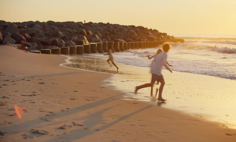 Three children laugh and play chase on the sandy shoreline of a beach as a golden sun sets over the ocean waves.