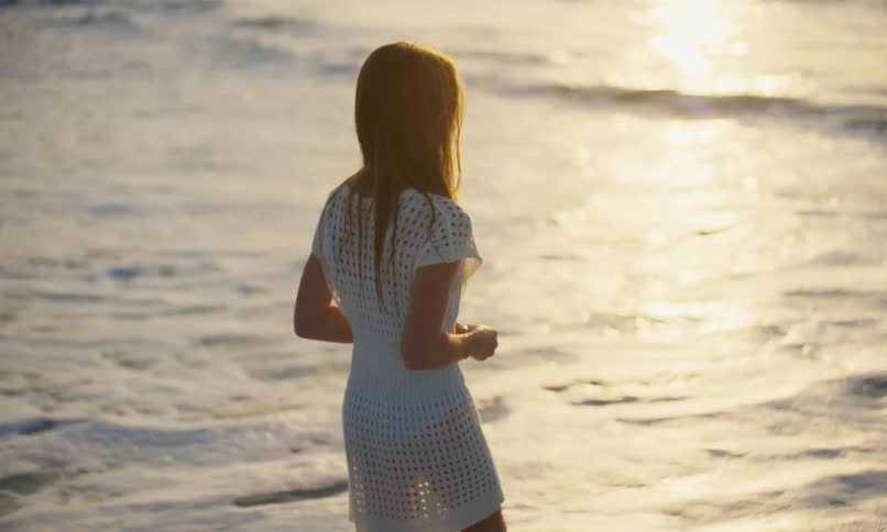 A young girl in a white swimsuit coverup dress is standing in the surf at the beach as the waves roll in and lifts her hand to shade her eyes from the setting sun.
