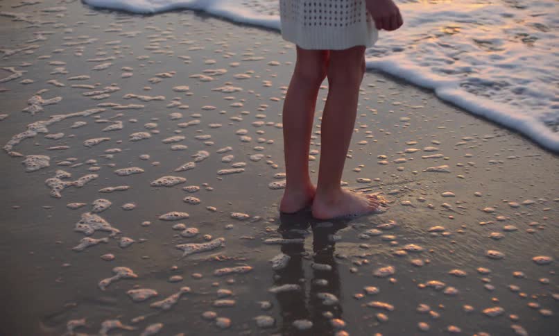 A young girl leaves footprints in the sand as she walks toward the wet shoreline of a beach at sunset and lets the incoming foamy surf swirl around her bare feet.