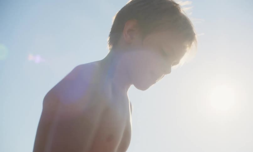 View from the sand looking up at a young, shirtless blonde boy as he digs a hole at the beach with a red plastic shovel with the sun shining down on him on a clear summer day.