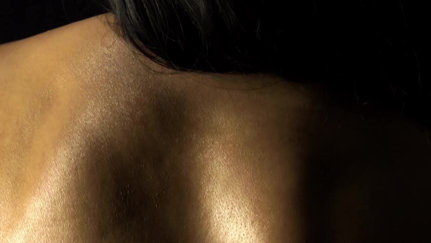 A closeup captures the toned smooth skin of a persons upper back and shoulders with dark hair cascading over the left shoulder against a black background