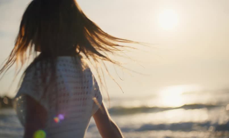 Slow-motion close-up of a young girl with long, damp hair and a white sundress happily twirling around with arms wide as she enjoys the sunset on an ocean beach.