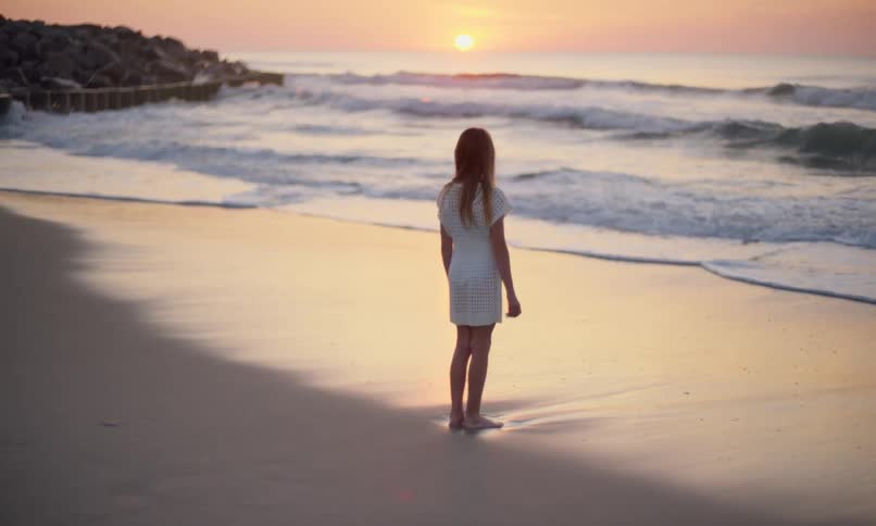 A young girl with long hair and a white dress stands alone on the beach peacefully watching the waves come in as the sun sets over the ocean.