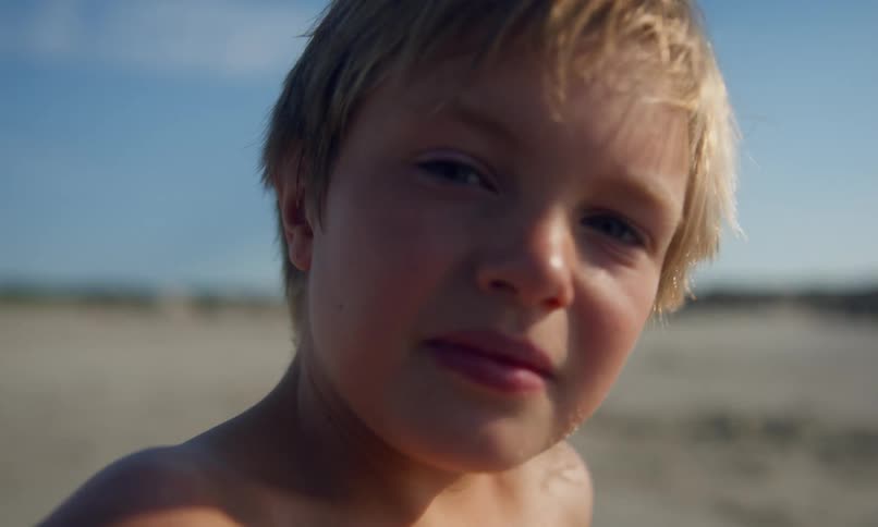 Close up of a young blonde boy sitting on a beach with his head turned away looking at something in the distance and then he turns to look at the camera and gives a cute smile.