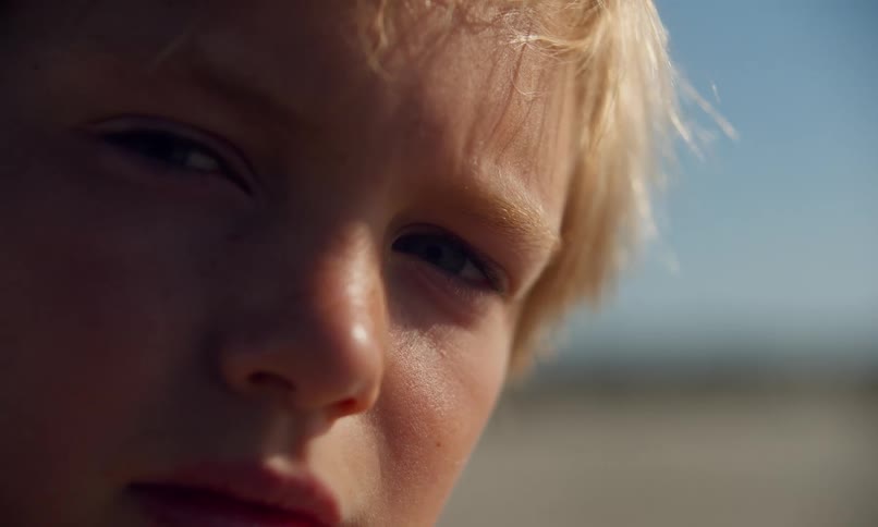 Close up of a young boy with blonde hair blowing in the breeze who looks directly into the camera and then away on a bright sunny day at the beach.