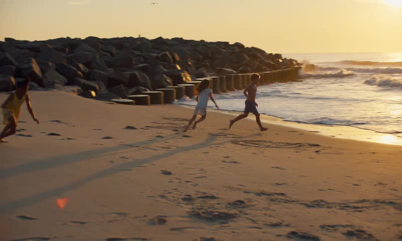 Three kids laugh, play, and make memories together on the shoreline of a sandy beach in front of a beautiful golden sunset.
