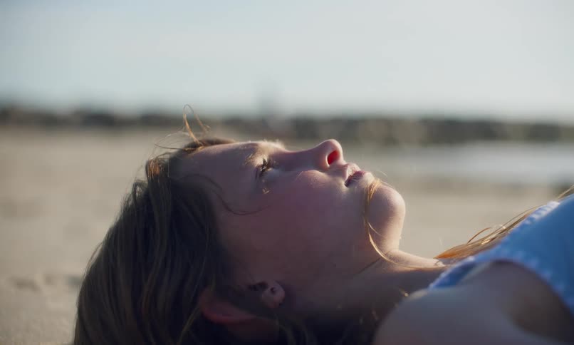A young girl lies basking in the sun on a sandy beach and then squints her eyes and sits up as blurred figures walk by in the background.