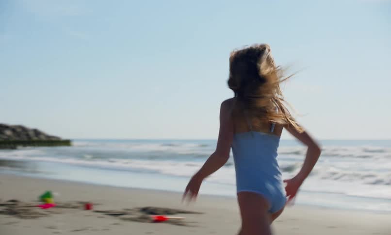 3 children, two girls and a boy, play together on a sandy beach on a sunny day, with two of them chasing each other, and another standing and shyly observing as the waves bring the surf to the shore.