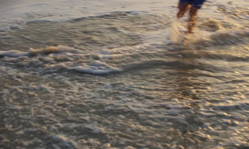 Close up of the surf coming in to a sandy beach shoreline with the rippled shadow reflections of 3 children playing and running from the waves as they wash up.