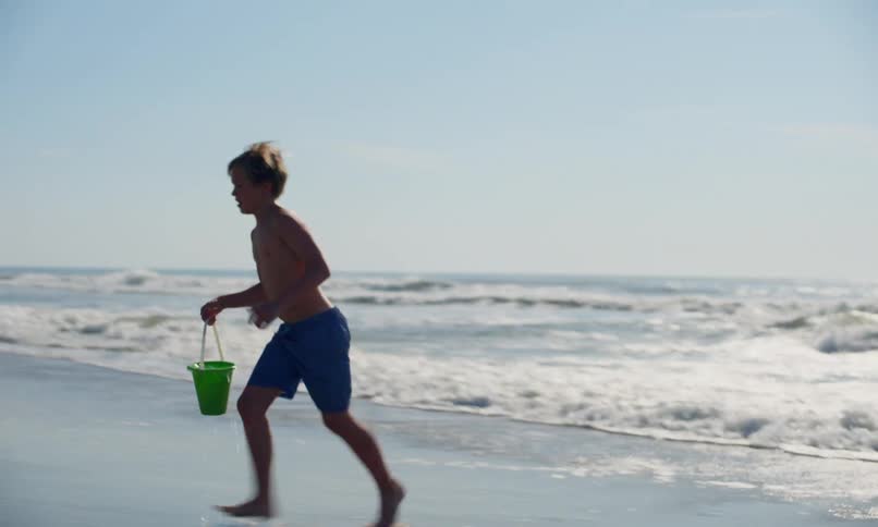 A young blonde boy wades into the surf at the beach and gathers water in a bucket to take back and pour into a hole where two girls are playing in the sand