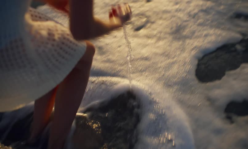 A young girl walking on the beach at sunset crouches to rinse a translucent piece of seaglass in the surf and then holds it up to the evening sunlight to look through it.