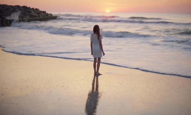 A young girl in a white dress stands deep in thought on the wet sandy shoreline of a beach peacefully looking out at waves coming in as the golden glow of the setting sun paints the sky with color.