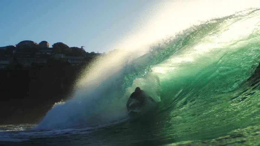 The surfer rides a huge wave by the homes on the tall cliff