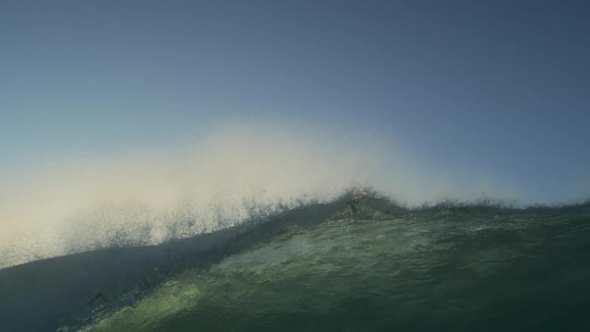 A large ocean wave is illuminated by sunlight under a pale blue sky at morning