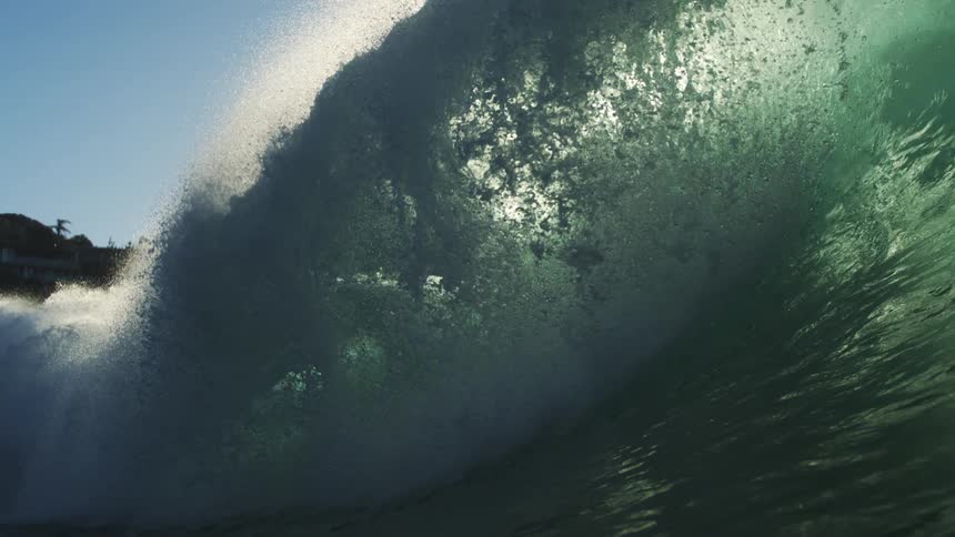 A large sun drenched ocean wave dramatically crests and barrels before the view abruptly transitions underwater into the turbulent green depths filled with light and bubbles