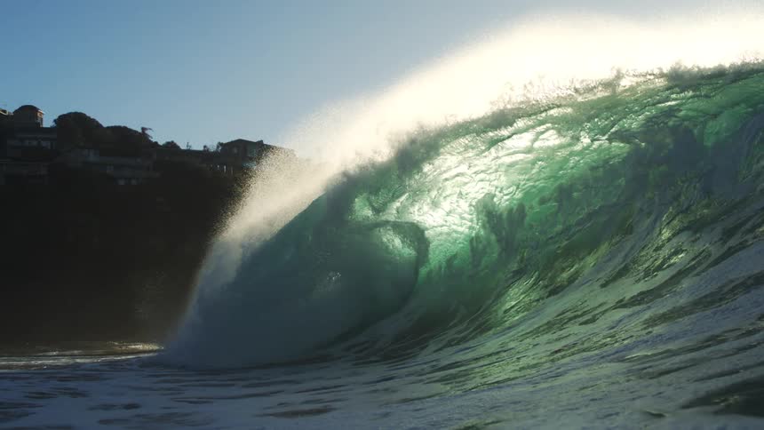 A surfer is captured midair off the face of a powerful wave as sunlight bursts over a cliffside