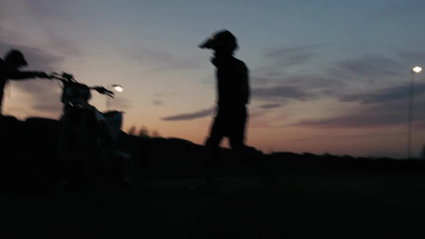 A man on helmet walks towards a motorcycle and climbs it at sunset