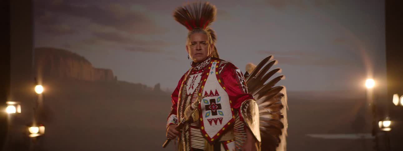 A man dressed in traditional native american attire including a feathered headdress and ornate clothing stands in a desert landscape with a distant rock formation