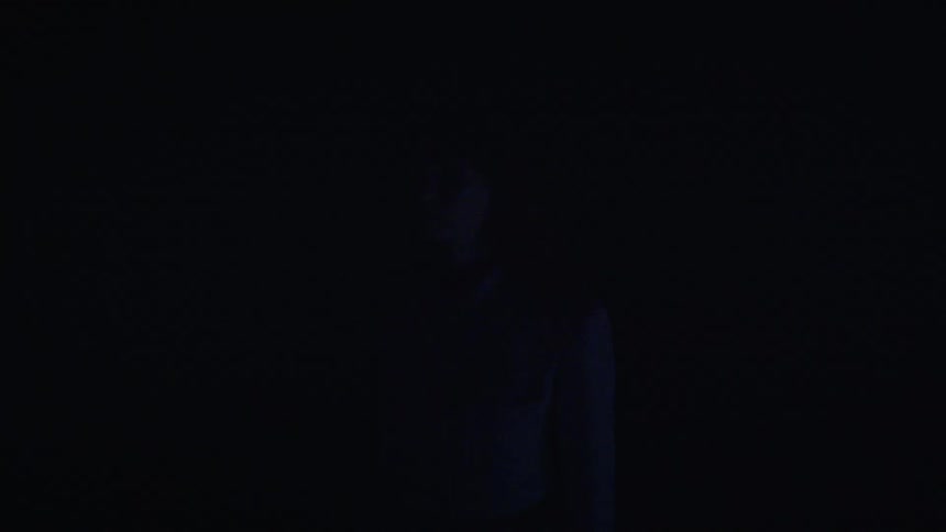 A young woman stands in a tunnel as automotive lights briefly illuminate the area revealing her before it goes dark again