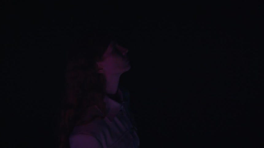 A female model with long hair stands in a dark room where light briefly illuminates her