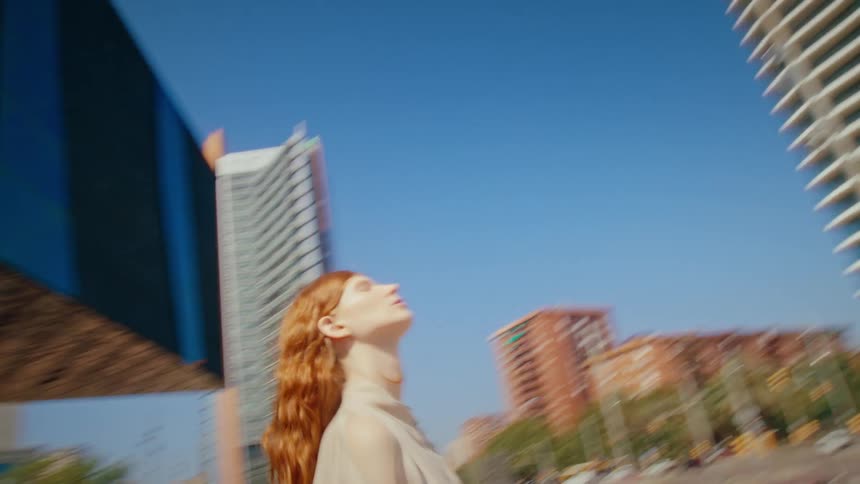 A blonde lady standing on a city road at midday stares at skyscraper buildings