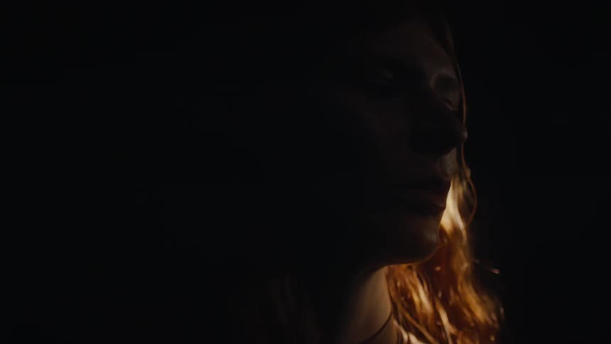 A woman with long light brown hair is partially visible in a dimly lit environment illuminated by a warm directional light that casts a soft glow on her features