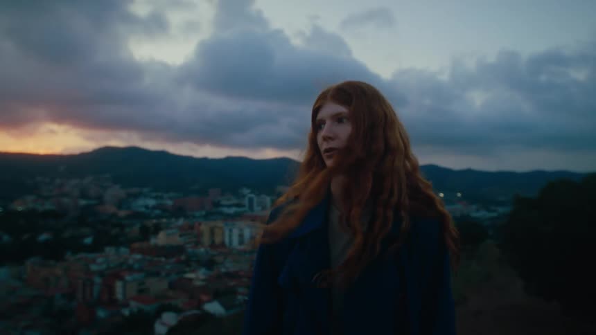 A woman with curly red hair is walking outside with her back turned and then she turns to look back and walks forward again