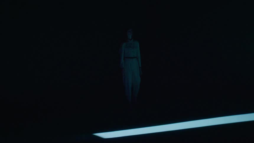 A lone woman walks down a runway into a bright rectangular spotlight that casts a long distinct shadow against a dark background