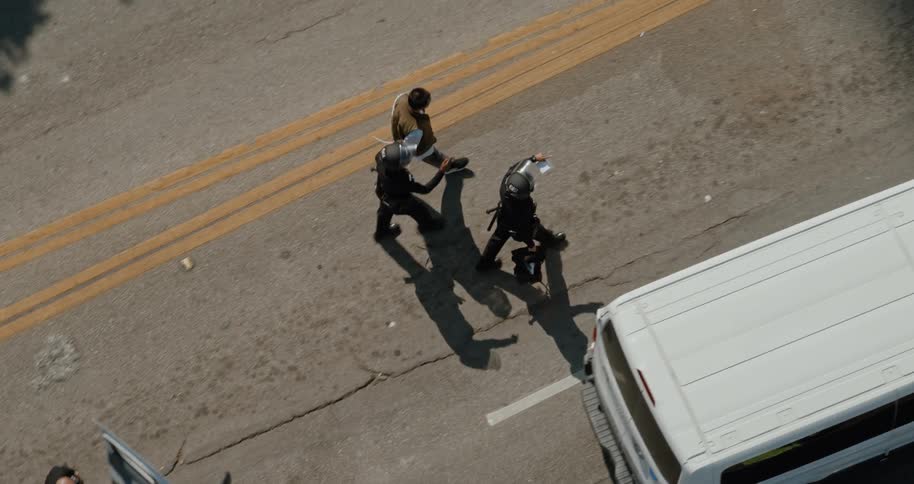 Three police officers escorting a handcuffed individual along a road with double yellow lines near a row of white vans