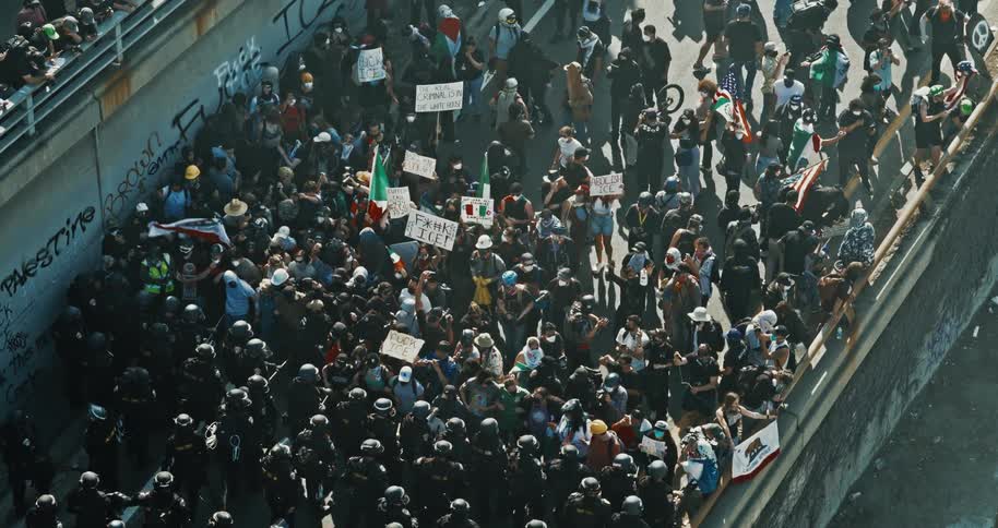 A large group of protesters holding signs and flags confronts a line of police officers in riot gear on a street next to a graffiticovered wall