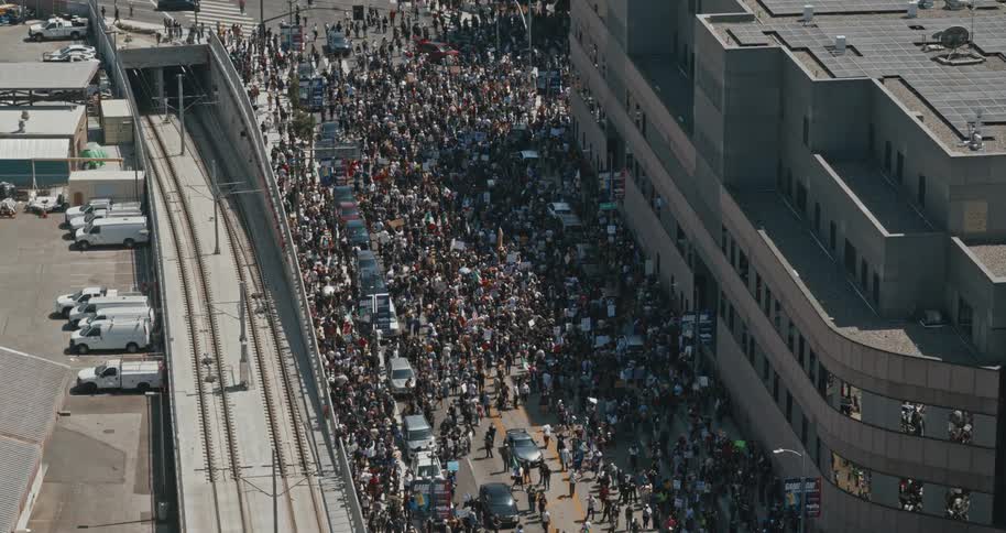 A large crowd of people is gathered on a street between a railway track and a building actively participating in a protest or demonstration