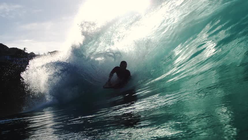 A lone bodyboarder rides inside a large curling ocean wave with bright sunlight and lens flares visible through the cresting water