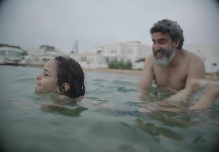 A child is swimming in the sea water of a beach next to an adult man who is helping