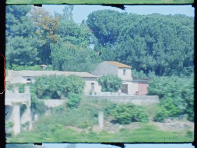 A sprawling house with a redtiled roof is nestled among green trees on a hill overlooking a vibrant green field