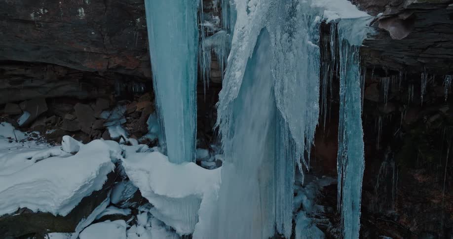 Water falls over the side of an ice covered rock cliff into a pool of water surrounded by snow covered rocks below