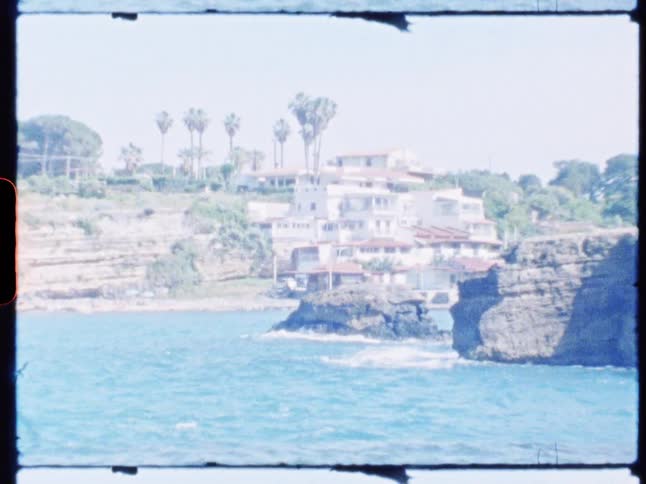 A coastal scene featuring rocky cliffs with waves crashing below surrounded by palm trees and greenery with a large white building perched nearby