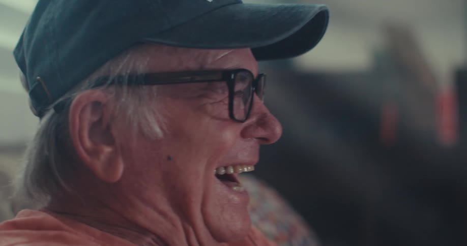An elderly man in glasses and a cap laughs joyfully in warm indoor light