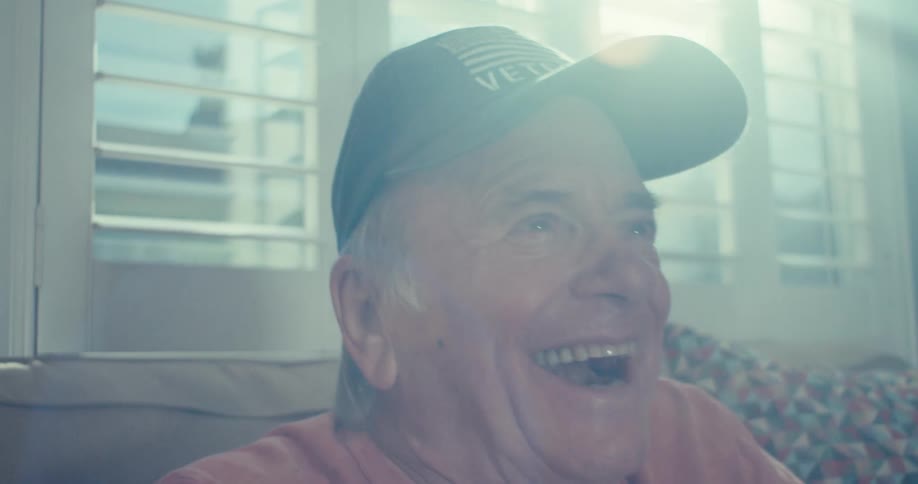 An elderly man in a cap smiles joyfully while seated indoors