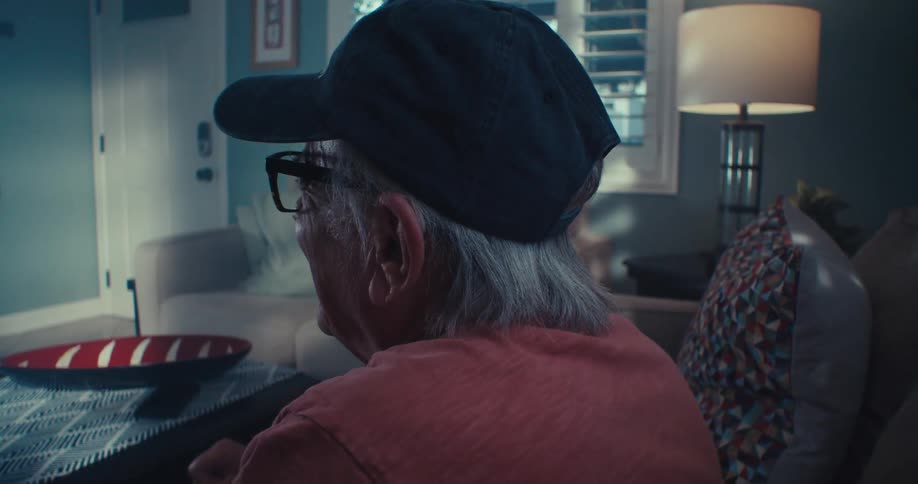Elderly man in black baseball hat sitting watching television