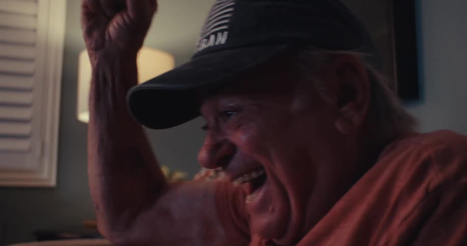 An older man smiles and pumps his fist in the air while excitedly while seated on a couch