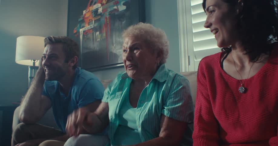 Three people sitting on a sofa and are happy looking forward and the lady in the center points forward