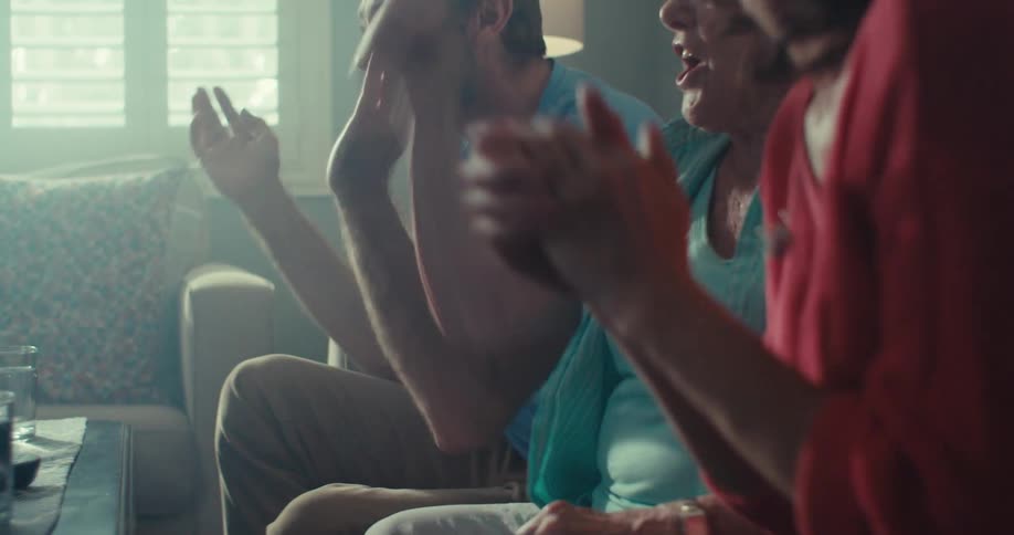 A couple sits on a couch with an elderly woman between them all excited and cheering while watching a tv show at midday